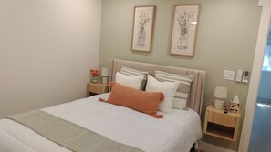 Een net opgemaakt bed met een witte dekbedovertrek en één oranje kussen, met aan weerszijden twee nachtkastjes met lampen en een klein bloemetje.