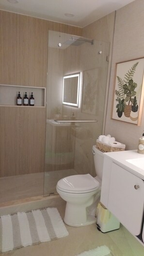 A modern bathroom with a glass-enclosed shower, a toilet, a sink, and a small shelf with toiletries. - Sunrise at Sarasota (Sarasota)
