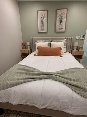 Een net opgemaakt bed met een witte dekbedovertrek en een groene gebreide plaid. Boven het bed hangen twee ingelijste foto's.
