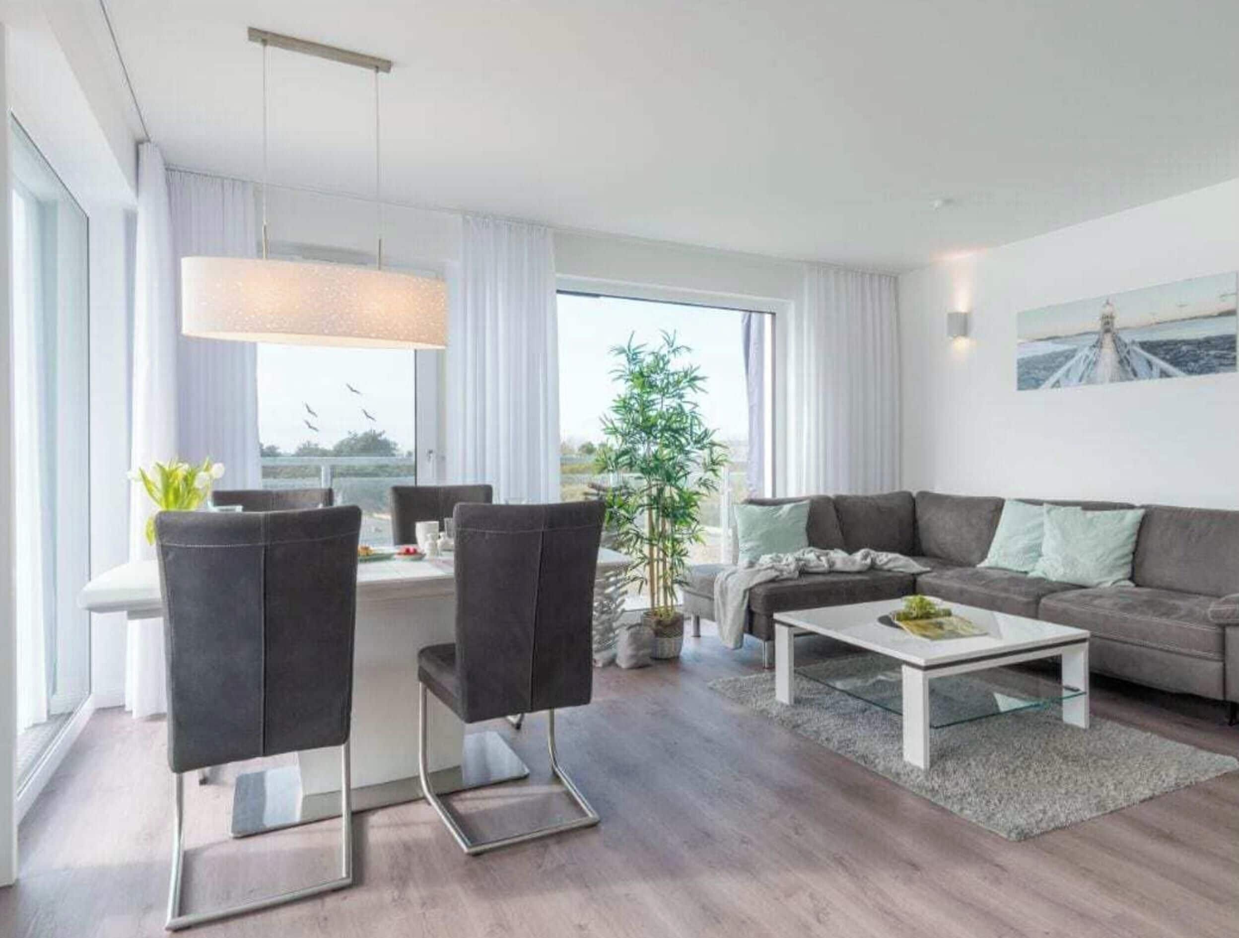 Un moderno salón con sofá, mesa de centro y sillas. Hay una zona de comedor con mesa y sillas. Un amplio ventanal ofrece vistas al exterior.