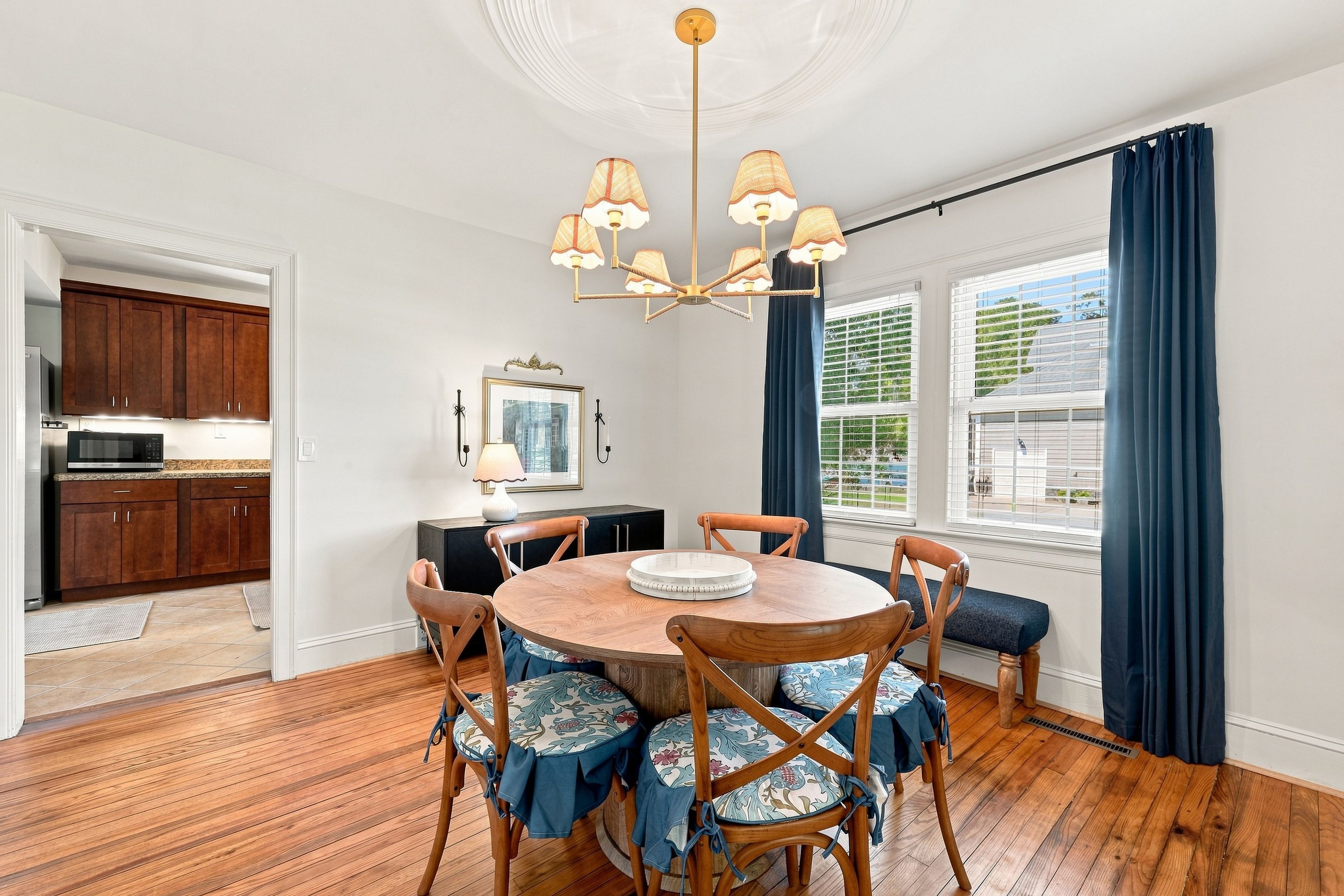 Un coin repas avec une table ronde en bois, des chaises à motifs floraux, un lustre et une vue sur l’extérieur grâce à de grandes fenêtres ornées de rideaux bleus.