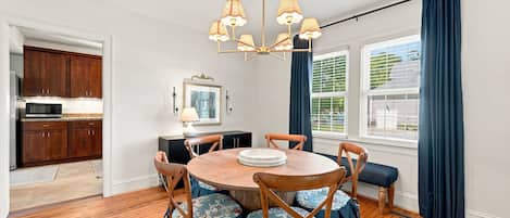 Un coin repas avec une table ronde en bois, des chaises à motifs floraux, un lustre et une vue sur l’extérieur grâce à de grandes fenêtres ornées de rideaux bleus.