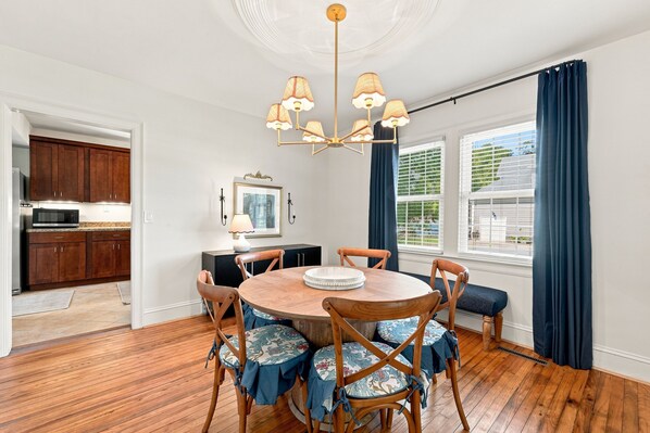 A dining area with a round wooden table, chairs with floral upholstery, a chandelier, and a view of the outdoors through large windows with blue curtains. - Cutest 3 BR cottage in New Bern close to everything! Welcome to the New Bernian! (New Bern)