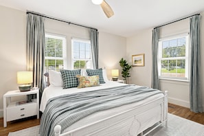 Un dormitorio con una cama, mesitas de noche, una lámpara, una planta y una ventana con cortinas.