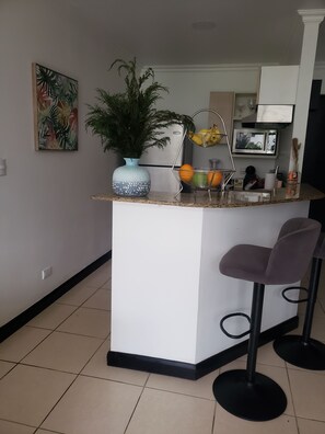 A kitchen area with a central island, bar stools, a potted plant, a fruit basket, and a microwave. - Oasis Apartment in the City at the Avalon Country Club Condominium in Costa Rica (Río Oro)