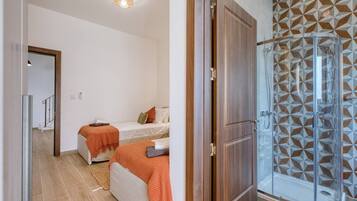 A bedroom with a bed, a bedside table, and a nightstand. There is a bathroom with a shower and a glass door.