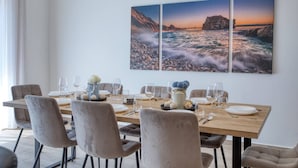 Un comedor con una mesa de madera dispuesta para seis personas, rodeada por seis sillas. Encima de la mesa hay tres cuadros enmarcados con paisajes costeros.