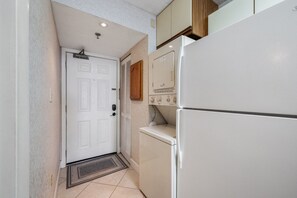 A compact kitchen with a white refrigerator, a washing machine, and a door with a vent. - San-A-Bel Oceanfront Getaway | 2BR (North Myrtle Beach)