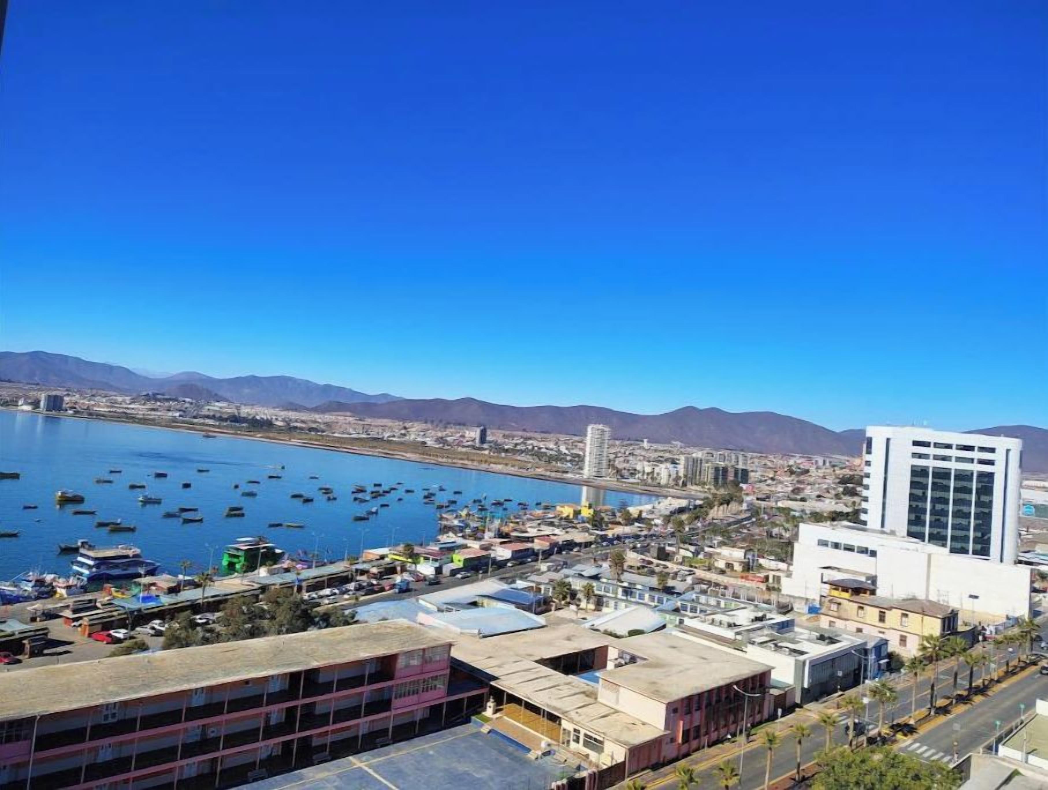 Un paysage urbain côtier avec un port de plaisance, des bâtiments et des montagnes en arrière-plan.