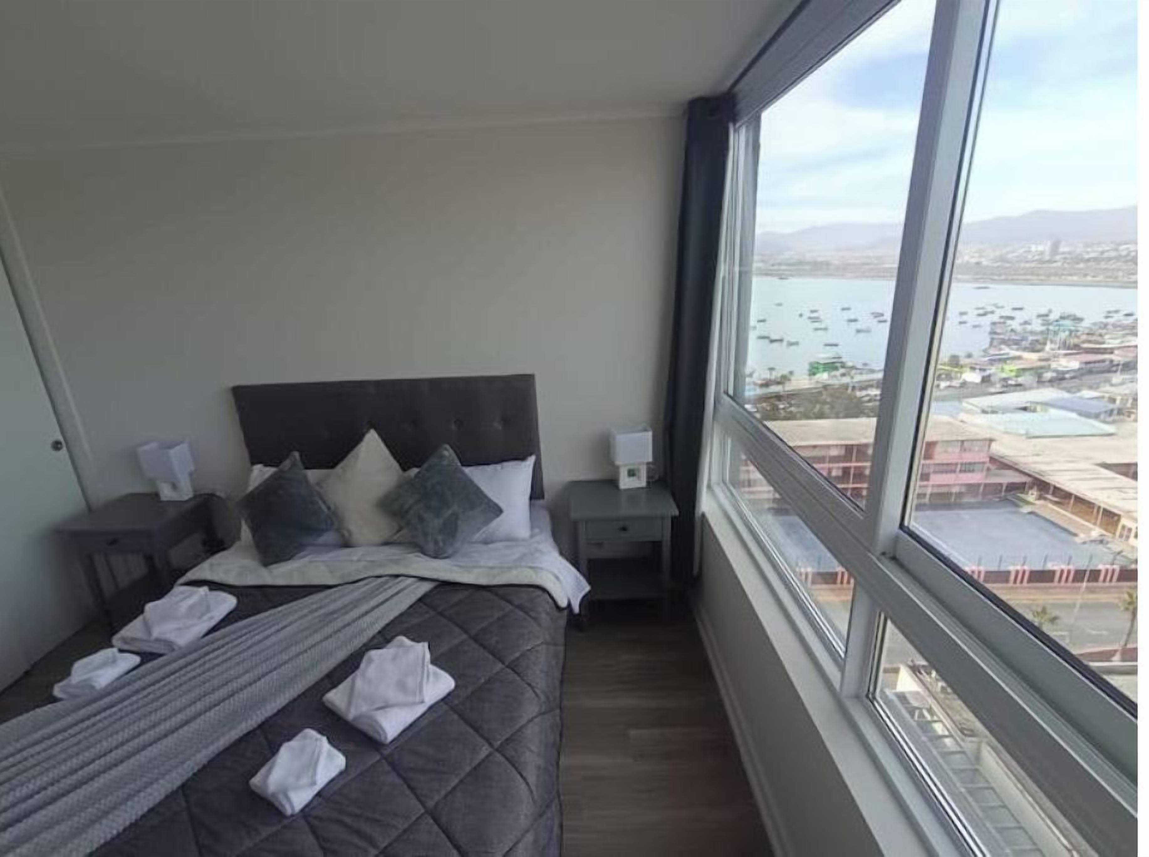 A hotel room with a large bed, a nightstand, a window with a city view, and a view of the water.