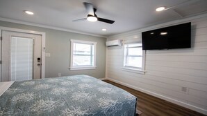 Ein Schlafzimmer mit einem Bett, einem Deckenventilator, einem Wandfernseher und zwei Fenstern.