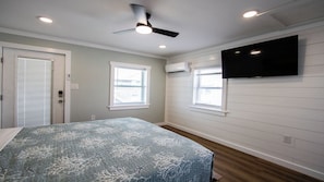 Une chambre à coucher avec un lit, un ventilateur de plafond, un téléviseur fixé au mur et deux fenêtres.