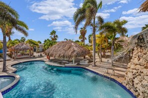 A swimming pool with a thatched-roof structure, surrounded by palm trees and a stone wall. - Casa Divers Dream - Screened porch and shared pool! (Kralendijk)