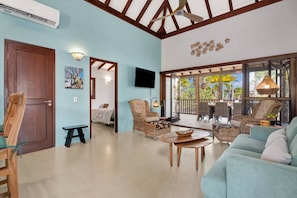 A spacious living area with a large sofa, a dining table, and wicker chairs. There is a television mounted on the wall, and a ceiling fan is visible. - Casa Divers Dream - Screened porch and shared pool! (Kralendijk)