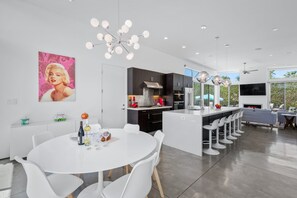 A modern kitchen with dark cabinetry, a central island, and a dining area with white chairs. - Flamingo Oasis | Private Pool & Spa | Firepit | Sleeps 6 (Palm Springs)