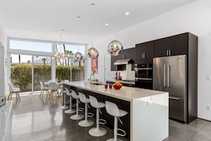 A modern kitchen with a central island, bar stools, stainless steel appliances, and large windows. - Flamingo Oasis | Private Pool & Spa | Firepit | Sleeps 6 (Palm Springs)