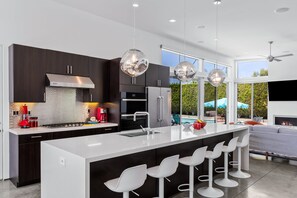 A modern kitchen with dark cabinetry, a large island with bar stools, and pendant lighting. - Flamingo Oasis | Private Pool & Spa | Firepit | Sleeps 6 (Palm Springs)