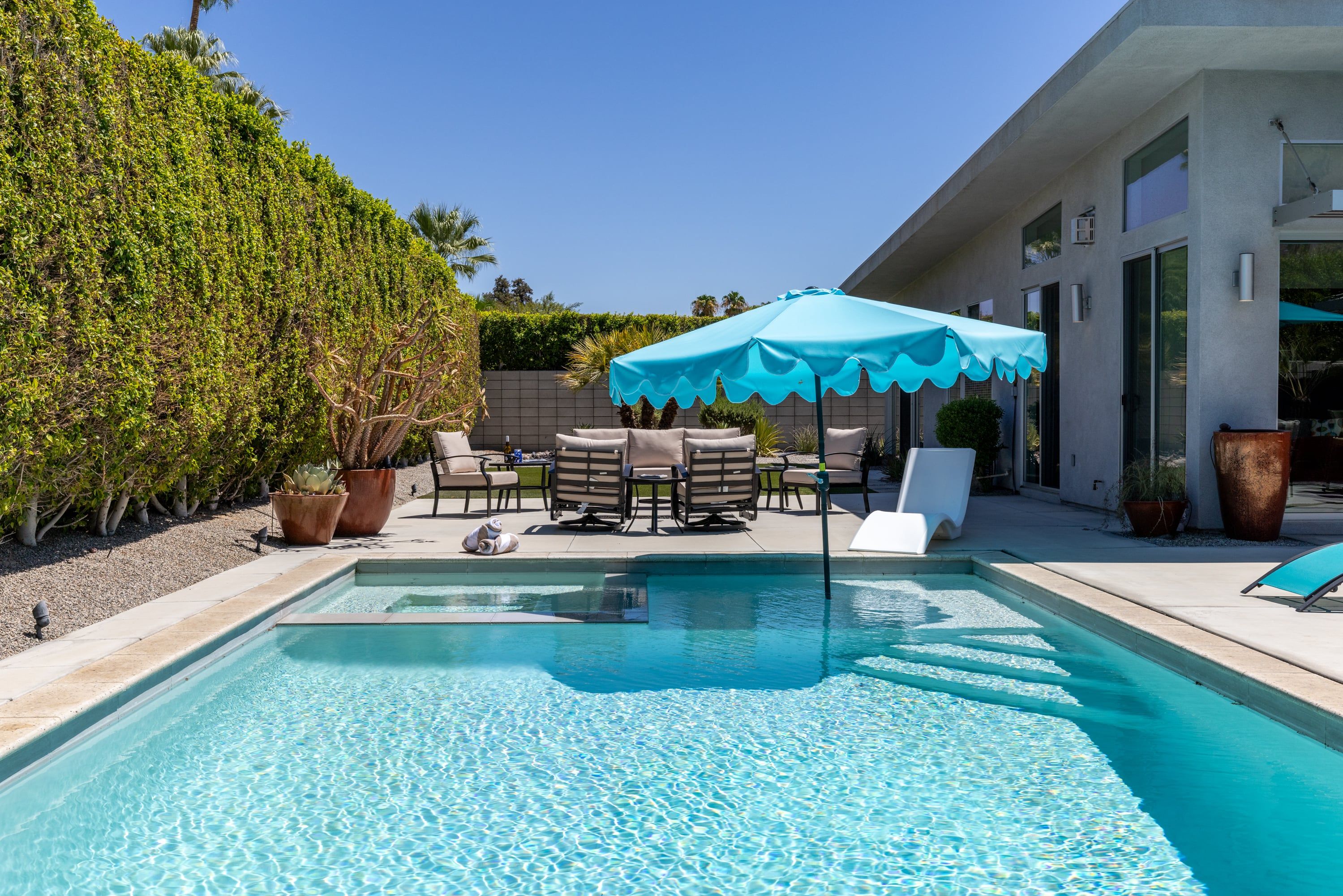 A swimming pool with lounge chairs and an umbrella, surrounded by a hedge and a building.