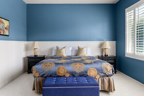 Ein Schlafzimmer mit blauer Wand, einem Bett mit gemustertem Überwurf, zwei Nachttischen mit Lampen und einem Fenster mit Jalousien.