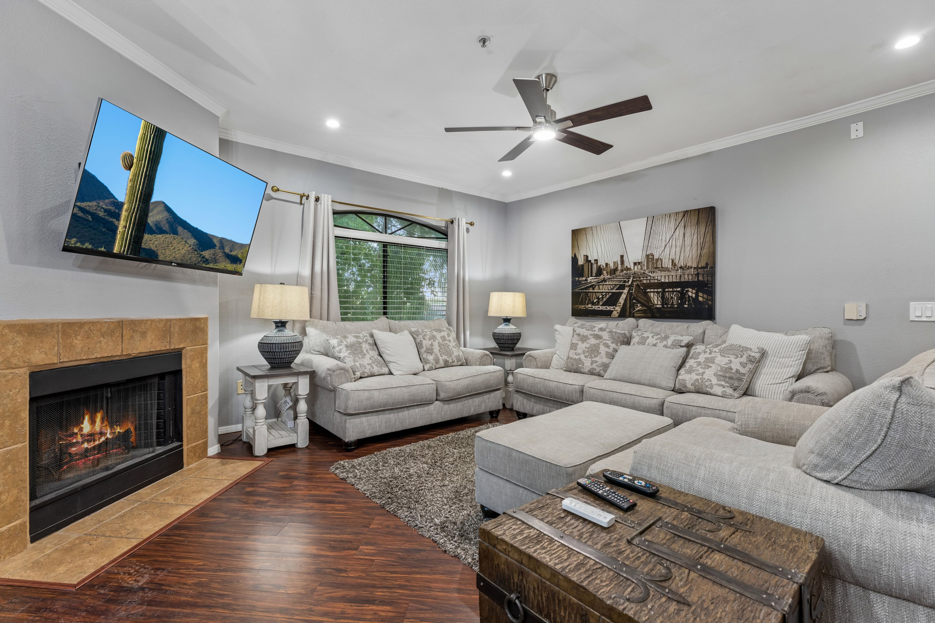 A living room with a fireplace, a ceiling fan, a large window with curtains, and a flat-screen TV mounted on the wall.