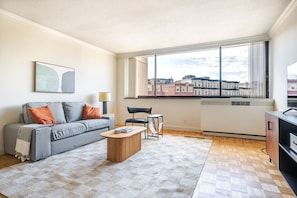 A modern living room with a gray sofa, a wooden coffee table, a black chair, and a large window with a city view. - Blueground | Fenway–Kenmore, gym, nr Fenway Park (Boston)