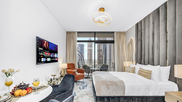 Ein modernes Hotelzimmer mit einem großen Bett, einem Schreibtisch mit einem Flachbildfernseher, einem Essbereich mit einer Obstplatte und Blick auf die Stadtlandschaft.
