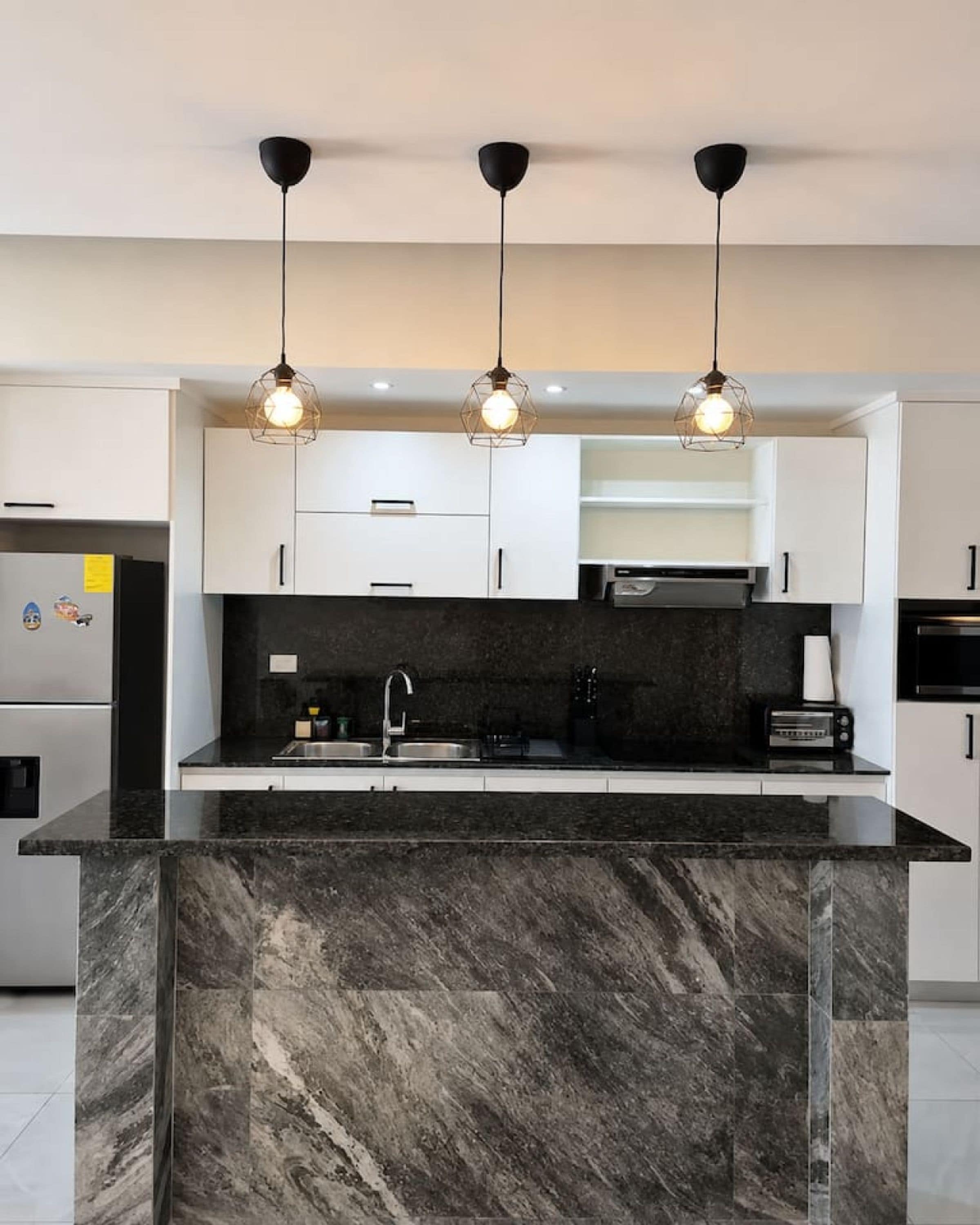 Cozinha moderna com ilha de mármore, bancadas pretas, armários brancos e iluminação pendente.