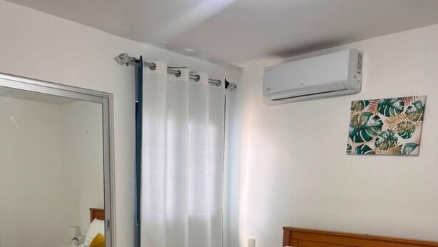 A bedroom with a wooden bed, white bedding, a mirror, a curtain, an air conditioning unit, and a wall-mounted lamp. - Relaxing 1BR Stay Gated Community (Punta Cana)