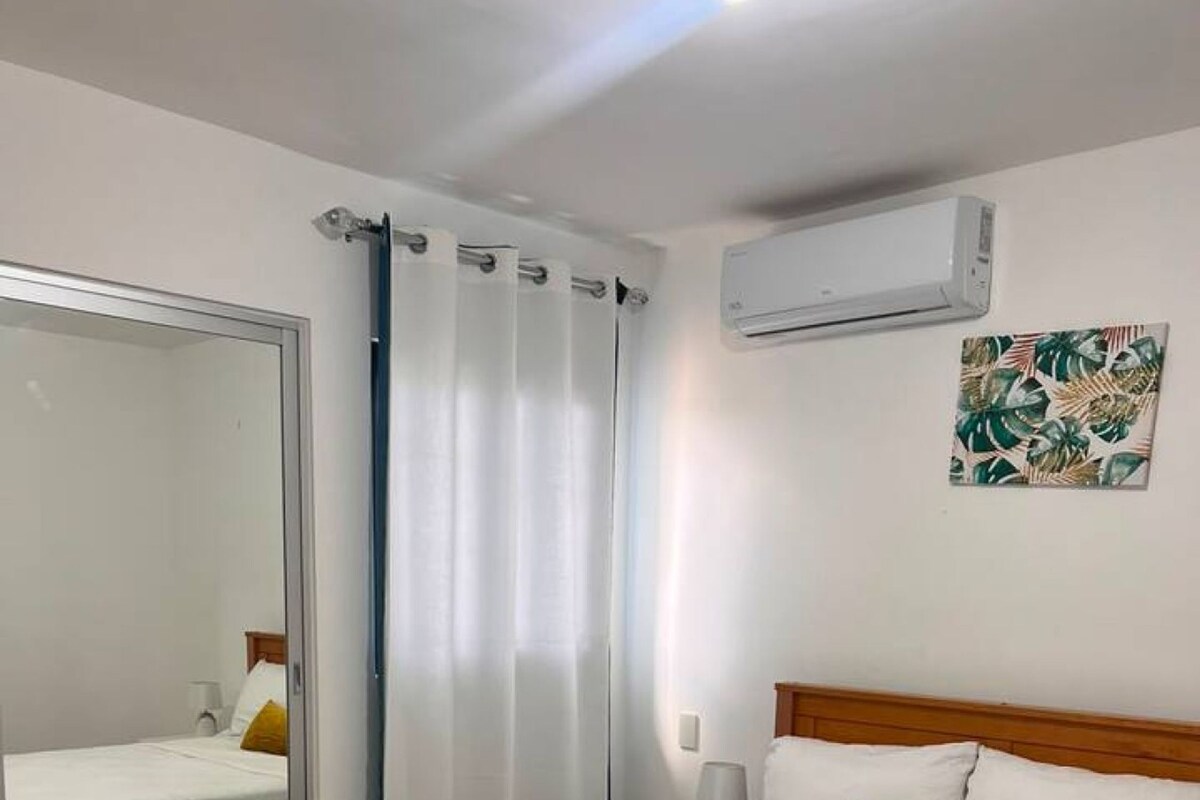 A bedroom with a wooden bed, white bedding, a mirror, a curtain, an air conditioning unit, and a wall-mounted lamp.