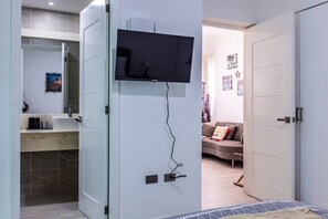 Quarto moderno com cama, TV fixada na parede, banheiro com pia e espelho, e sala de estar com sofá e decorações.