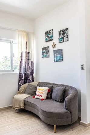 A modern living room with a grey sofa, a window with curtains, and cityscape artwork on the wall. - Cozy 1BR Beach Pool Electricity Included (Punta Cana)