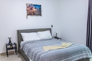 Quarto com cama, mesa de cabeceira, abajur, quadro na parede e cortina.