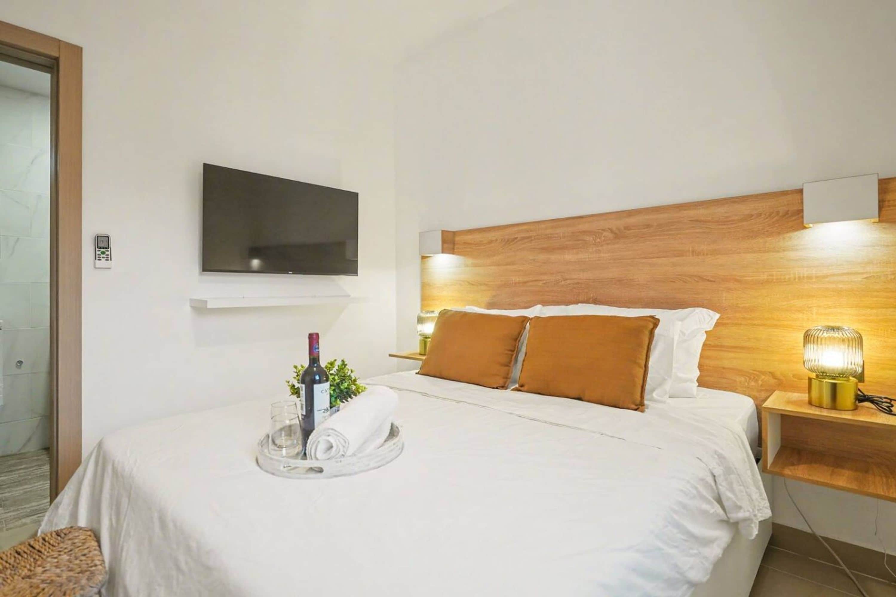 Una habitación de hotel con una cama, un televisor montado en la pared, una mesita de noche con una lámpara y una bandeja con una botella y vasos.