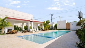 Una piscina con sillas de descanso y una zona de estar techada, rodeada de vegetación y un edificio blanco.