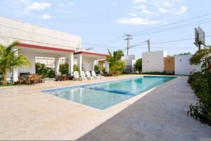 Piscina com espreguiçadeiras e área de estar coberta, cercada por vegetação e um edifício branco.
