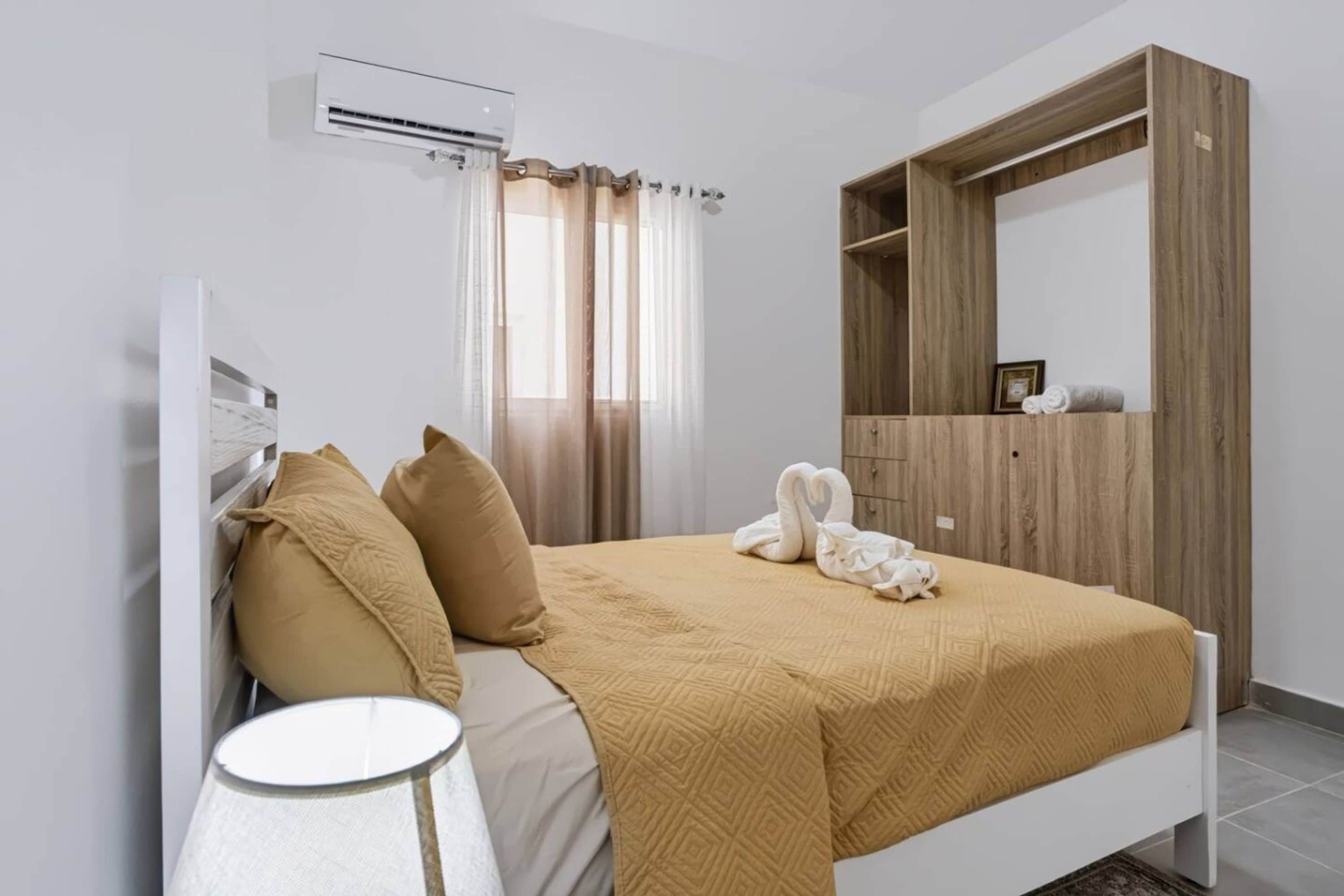 Un dormitorio con cama, mesita de noche, aire acondicionado, armario y ventana con cortinas.