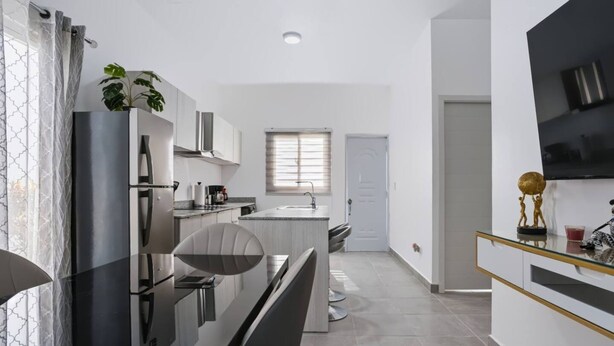 Una cocina moderna con una isla central, un comedor con una silla negra y un televisor montado en la pared.
