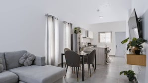Una sala moderna con un sofá gris, una mesa de comedor con sillas, una cocina con refrigerador y una pequeña mesa de comedor con una planta.