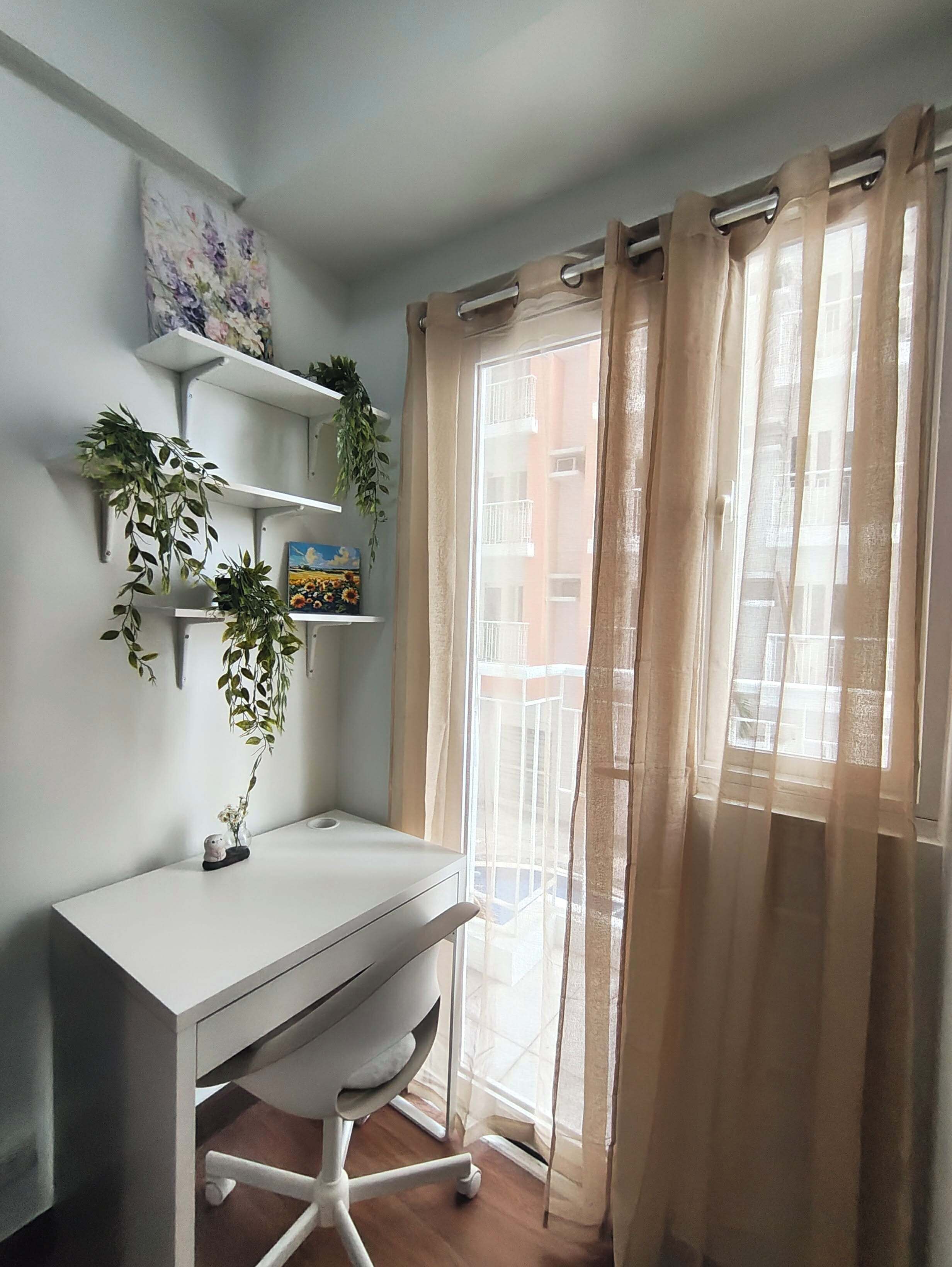 Un espacio de oficina moderno con un escritorio blanco, estanterías con plantas y vistas a través de una ventana con cortinas translúcidas.