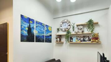 A modern living room with a sofa, decorative pillows, wall art, a clock, shelves with plants and books, and a TV.