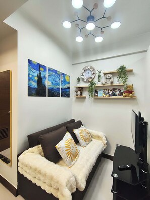 A modern living room with a sofa, decorative pillows, wall art, a clock, shelves with plants and books, and a TV.