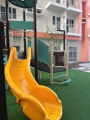 A playground with a yellow slide and climbing structures, set against a building with balconies and a tree.