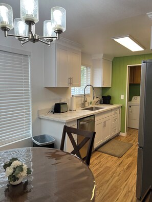 A kitchen with white cabinets, a stainless steel refrigerator, a sink, and a dining table with a flower vase. - Sequim and Sun and Fun (Sequim)