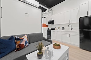 A modern kitchen with white cabinets, a black refrigerator, and a grey sofa with colorful pillows. - Dine, Shop & Explore | Walk to Petco Park & More!! (San Diego)