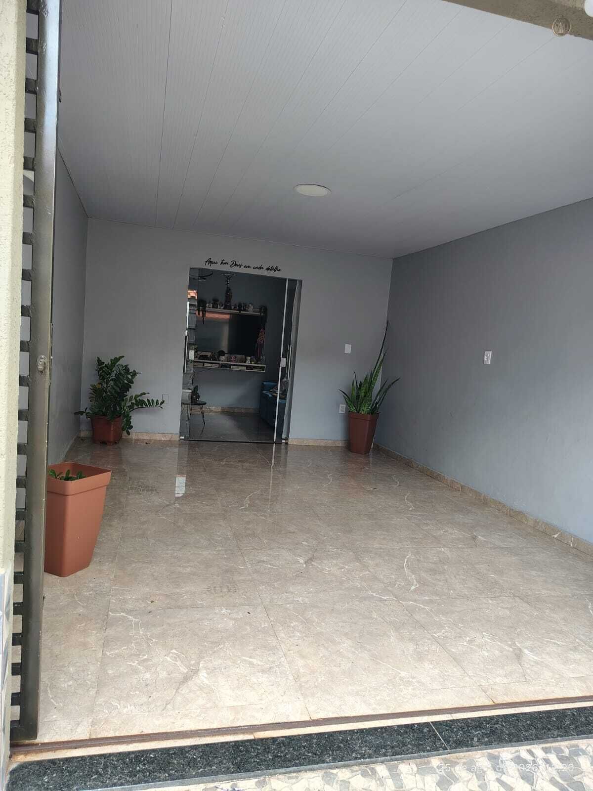 Un espace intérieur spacieux doté d’un sol carrelé, de deux plantes en pot, d’une porte-fenêtre donnant sur une pièce avec une étagère intégrée et d’un luminaire au plafond.