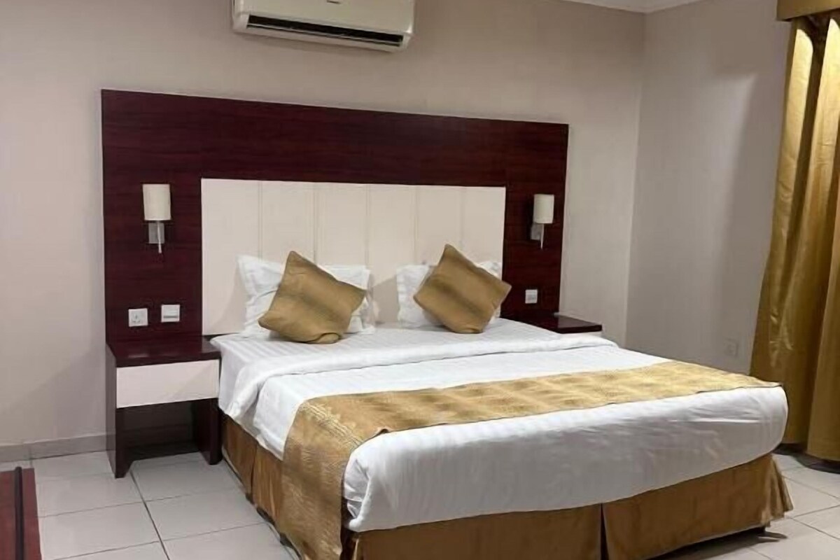 A hotel room with a large bed, a headboard, two bedside tables, an air conditioning unit, and a curtain.