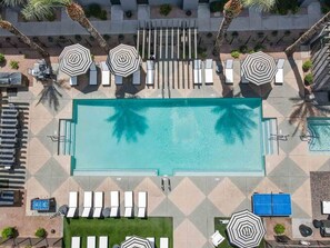 An aerial view of a swimming pool area with multiple lounge chairs and umbrellas, surrounded by palm trees and a tiled deck. - CozySuites Glendale - Westgate w pool! 2BR 2BA (Glendale)