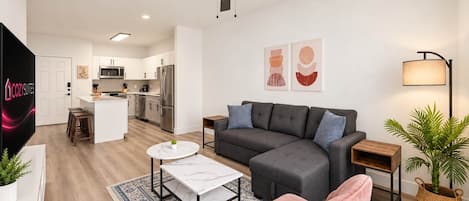 Ein modernes Wohnzimmer mit einem grauen Sofa, einem Couchtisch und einem rosafarbenen Sessel. Im Hintergrund ist eine Küche mit weißen Schränken und Edelstahlgeräten zu sehen.