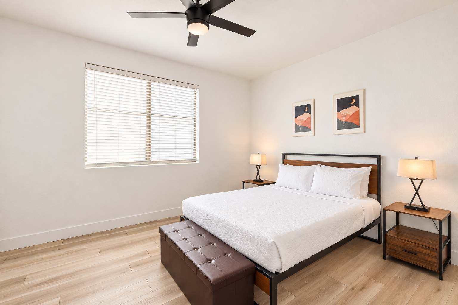 A bedroom with a bed, two bedside tables, a ceiling fan, a window with blinds, and two wall-mounted artworks.