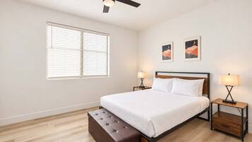 A bedroom with a bed, two bedside tables, a ceiling fan, a window with blinds, and two wall-mounted artworks.